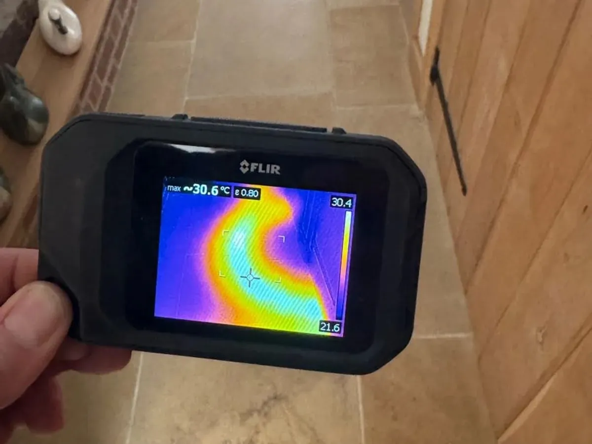 FLIR thermal camera held over a tiled hallway, its screen showing a warm curving underfloor heating pipe